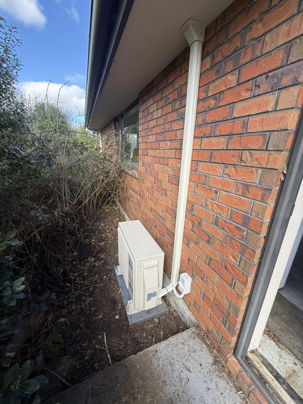 Heat pump outdoor unit installed on a pad beside a brick house, with tidy cover over the pipework